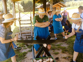 Eine Gruppe von Menschen, die Hüte tragen und Schürzen anhaben, arbeitet gemeinsam an einem Holzprojekt im Freien. Sie nutzen eine große Säge, um Holzstücke zu schneiden, während andere in der Nähe zuschauen. Im Hintergrund sind Bäume und eine Hütte sichtbar.