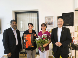 Vier Personen halten ein Geschenk und einen Blumenstrauß in einem feierlichen Moment.