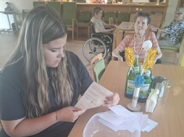 Eine junge Frau liest eine Broschüre an einem Tisch mit Seniorinnen im Hintergrund.