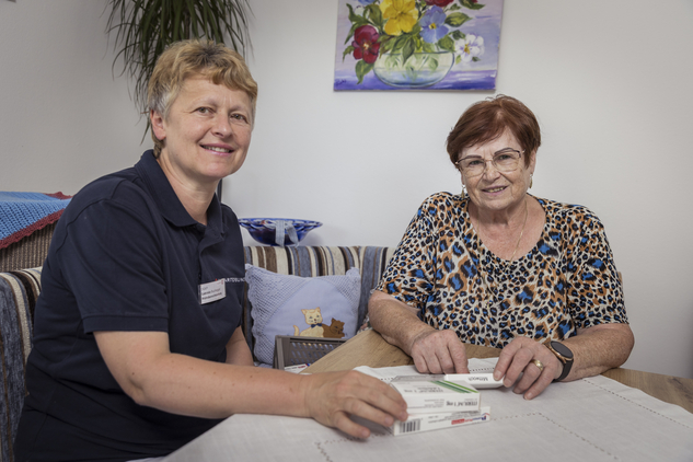 Eine Pflegerin und eine ältere Frau sitzen am Tisch mit Medikamenten.