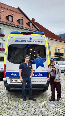 Zwei Männer stehen neben einem Wagen mit der Aufschrift „Samariter-Wunschfahrt“.