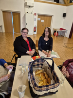 Zwei Frauen sitzen an einem Tisch in einem Raum. Auf dem Tisch steht ein Korb mit kleinen Brötchen. Im Hintergrund sind Türen und einige Gegenstände zu sehen. Es herrscht eine gemütliche Atmosphäre.