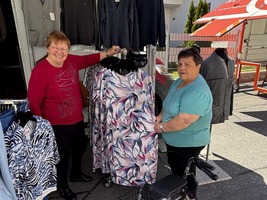Zwei Frauen stehen in einem Bekleidungsstand. Eine Frau hält ein Kleidungsstück mit einem blühenden Muster, während die andere daneben steht und lächelt. Im Hintergrund sind weitere Kleidungsstücke auf Kleiderstangen zu sehen. Es ist ein sonniger Tag.