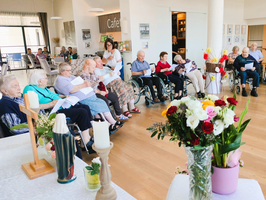 Eine Gruppe älterer Menschen sitzt in einem Gemeinschaftsraum auf Stühlen und Rollstühlen. Einige halten Blätter in den Händen. Im Vordergrund stehen Blumenarrangements und eine Statue. Der Raum wirkt einladend und hell.