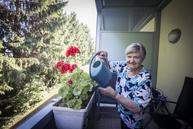 Eine ältere Frau gießt rote Geranien auf ihrem Balkon.