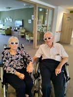 Ein älteres Paar in Rollstühlen trägt Sonnenbrillen und hält sich an den Händen.