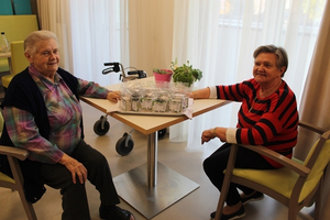 Zwei ältere Frauen sitzen an einem Tisch mit Pflanzen und Töpfen.