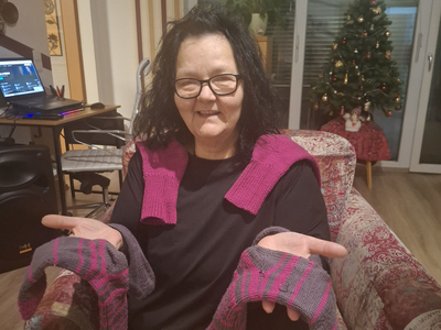 Eine Frau in einem Sessel zeigt gestrickte Handschuhe und einen Schal.