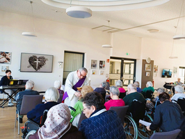 Gottesdienst mit Senioren, Pfarrer segnet und Musiker spielt im Hintergrund.
