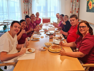 Eine Gruppe von zehn Personen in medizinischen Uniformen sitzt an einem Tisch und genießt eine Mahlzeit. Sie lächeln und halten Besteck in der Hand. Der Tisch ist mit verschiedenen Gerichten und Getränken gedeckt. Die Umgebung ist hell und freundlich, mit Pflanzen und Dekorationen.