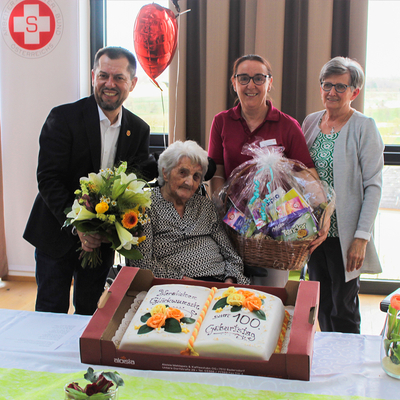 Eine ältere Frau sitzt lächelnd im Rollstuhl, umgeben von drei Personen. Sie hält einen Blumenstrauß, während eine zweite Person einen Geschenkkorb präsentiert. Vor ihnen steht eine große Geburtstagstorte mit der Aufschrift „Zum 100. Geburtstag“ und orangefarbenen Rosen.