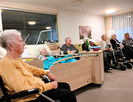 Sechs ältere Frauen sitzen in einem Wohnraum, eine Frau liegt auf einem Bett.
