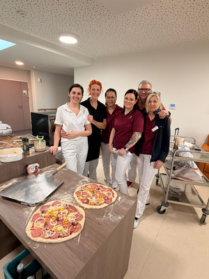 Sechs Personen in einem Raum stehen um einen Tisch mit zwei belegten Pizzen. Sie lächeln und posieren für das Foto. Im Hintergrund sind Küchenelemente und Zutaten zur Zubereitung von Pizza sichtbar.
