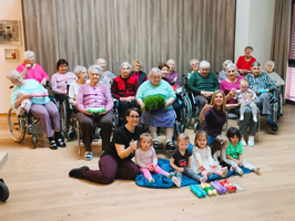 Eine Gruppe von älteren Menschen in Rollstühlen sitzt zusammen in einem Raum. Vor ihnen stehen mehrere kleine Kinder, die mit bunten Objekten spielen. Einige Erwachsene lächeln und interagieren mit den Kindern, während die Gruppe freundlich wirkt.