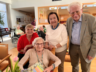 Eine Gruppe von vier älteren Menschen in einem Wohnheim. Drei stehen und lächeln, während eine Frau im Sessel sitzt. Auf dem Tisch sind Blumen und ein Notizbuch sichtbar. Im Hintergrund sind weitere Bewohner zu sehen.
