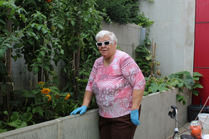Eine ältere Frau steht neben einem Gemüsegarten und trägt Sonnenbrille und Handschuhe.