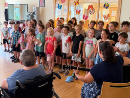 Eine Gruppe von Kindern steht in einem Raum und singt. Im Vordergrund sitzt eine Person im Rollstuhl und hört zu. Einige Kinder tragen bunte Kleidung, und im Hintergrund sind Schmetterlingsdekorationen an den Wänden zu sehen. Eine Erwachsene spielt Gitarre.