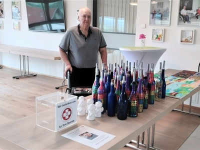 Ein älterer Mann steht neben bunten Flaschen auf einem Tisch.