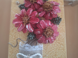 Ein dekoratives Handwerkstück zeigt eine florale Gestaltung aus roten Blumen, die auf einer gelb gemusterten Unterlage angeordnet sind. Der Blumenstrauß ist mit grünen Blättern und einer kleinen, dekorativen Schleife aus Jute umrahmt.