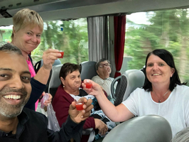 Eine Gruppe von fünf Personen in einem Bus hebt kleine Gläser an, während sie miteinander anstoßen. Im Hintergrund sind Bäume sichtbar, die an einem Fenster vorbeiziehen. Alle Personen wirken fröhlich und genießen den Moment.