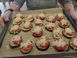 Eine ältere Frau hält ein Blech mit belegten Brötchen in einem Gemeinschaftsraum.
