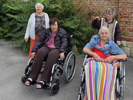 Vier ältere Frauen in Rollstühlen vor einer Backsteinmauer, umgeben von Pflanzen.