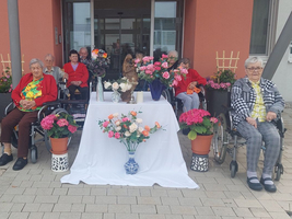 Eine Gruppe älterer Menschen sitzt draußen vor einer dekorierten Tischfläche mit Blumen.