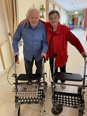 Ein älteres Paar steht in einem Flur, beide nutzen Rollatoren zur Unterstützung.