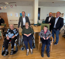 Eine Gruppe von vier älteren Frauen sitzt und steht in einem Raum. Zwei Frauen sitzen in Rollstühlen, während die anderen zwei auf Stühlen sitzen. Im Hintergrund stehen drei Männer in Anzügen. Der Raum sieht einladend aus, mit Tischen und Pflanzen.