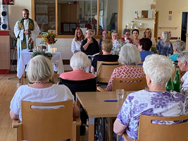 Ein Mann in Priesterkleidung spricht zu einer Gruppe älterer Frauen in einem Gemeinschaftsraum.