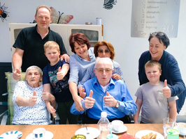Eine fröhliche Gruppe von sieben Personen posiert für ein Foto, darunter zwei ältere Menschen, die in Stühlen sitzen, und fünf jüngere Personen, die um sie herum stehen. Alle zeigen Daumen hoch und lächeln, um Glück und Zusammengehörigkeit auszudrücken. Im Vordergrund sind Teller und Getränke auf einem Tisch zu sehen.