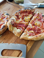 Eine frisch gebackene Pizza wird auf einem Holztisch präsentiert. Die Pizza hat eine knusprige Kruste und ist mit Tomatensauce, Schinken, Mais und Zwiebeln belegt. Ein Stück wurde bereits herausgeschnitten. Im Hintergrund sind einige Küchenutensilien sichtbar.