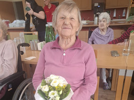 Eine ältere Frau sitzt auf einem Stuhl und hält einen Blumenstrauß mit weißen und gelben Blumen in der Hand. Im Hintergrund sind weitere Personen und ein Raum mit Tischen und Stühlen sichtbar.