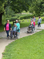 Mehrere Personen spazieren auf einem Weg in einem Park. Zwei Frauen schieben Rollstühle mit älteren Menschen, während mehrere andere Personen im Hintergrund in verschiedene Richtungen gehen. Die Umgebung ist grün und einladend, mit Bäumen und Rasen.