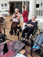 Zwei Frauen in Rollstühlen interagieren mit einer Pflegekraft und einem kleinen Welpen.