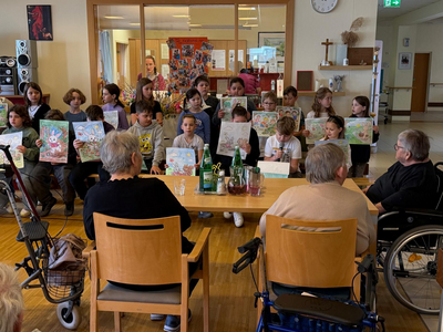 Eine Gruppe von Kindern steht in einem Raum und hält gemalte Bilder hoch. Vor ihnen sitzen zwei ältere Personen, während andere Personen im Hintergrund stehen. Der Raum wirkt einladend und ist mit verschiedenen Kunstwerken dekoriert.