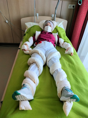 Person mit Verbänden liegt auf einem Bett in einem Krankenhauszimmer.
