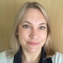 Porträt einer lächelnden Frau mit blonden, schulterlangen Haaren, die in einem weißen Oberteil und einem dunklen Poloshirt gekleidet ist. Der Hintergrund besteht aus einem hellen Holzregal.