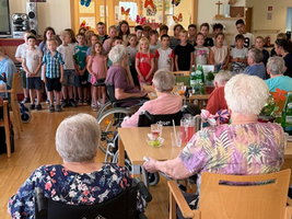 Eine Gruppe von Kindern steht in einem Raum und singt für ältere Menschen, die in Rollstühlen sitzen. Die Senioren sind am Tisch versammelt und beobachten die Darbietung. Die Umgebung ist freundlich und festlich dekoriert.