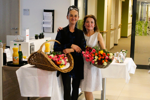Zwei Frauen stehen lächelnd mit Blumen und Getränken in einem Raum.