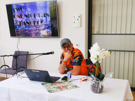 Ein Mann sitzt an einem Tisch mit einem Laptop, Mikrofon und einer Pflanze, während im Hintergrund ein Bildschirm mit der Aufschrift „TWO X ONE NIGHT IN BANGKOK“ zu sehen ist. Der Tisch ist mit einer dekorativen Tischdecke und einer Blume geschmückt.