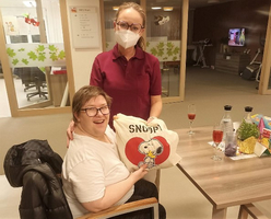Frau sitzt lächelnd mit einer Tasche und Krankenschwester im Hintergrund.