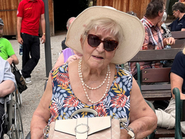 Eine ältere Frau in einem farbenfrohen Blumenkleid sitzt im Rollstuhl. Sie trägt eine große Sonnenbrille und einen breiten Sonnenhut und hält eine Handtasche sowie eine Tasse. Im Hintergrund sind andere Personen zu sehen, die sich im Freien aufhalten.