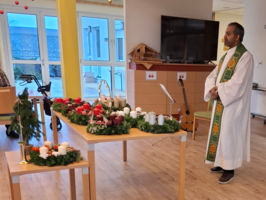 Ein Priester steht neben einem Tisch mit festlich dekorierten Kerzen und Pflanzen.