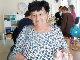 Eine ältere Frau mit schwarzem, lockigem Haar trägt ein gepunktetes Oberteil und hält ein Holzbrett mit bunten, dekorativen Gegenständen in der Hand. Im Hintergrund sind weitere Personen und ein Raum mit Tisch und Dekorationen zu sehen.