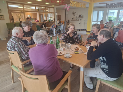 Eine Gruppe von sieben älteren Menschen sitzt an einem Tisch in einem Gemeinschaftsraum. Sie unterhalten sich und genießen Getränke und Kuchen. Im Hintergrund sind weitere Personen und Einrichtungen zu sehen, die eine freundliche, einladende Atmosphäre schaffen.