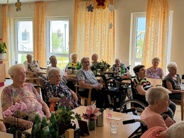 Eine Gruppe älterer Frauen sitzt in einem hellen Raum mit großen Fenstern. Einige Frauen sind in Rollstühlen, während andere auf Stühlen sitzen. Blumen und Pflanzen sind auf den Tischen verteilt, und die Umgebung wirkt freundlich und einladend.