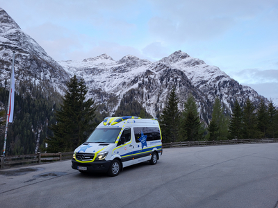 Ein Krankenwagen steht auf einer asphaltierten Straße vor schneebedeckten Bergen und einem bewaldeten Hang. Der Himmel ist bewölkt und die Landschaft vermittelt eine ruhige Bergatmosphäre.
