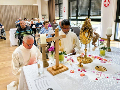 Ein Andachtsraum mit einer weißen Tischdecke, auf der ein Holzkreuz, Kerzen, Blumen und ein Monstranz stehen. Zwei Priester sitzen davor, einer betet und der andere liest. Im Hintergrund sind ältere Menschen aus einer Gemeinde versammelt, einige im Rollstuhl.