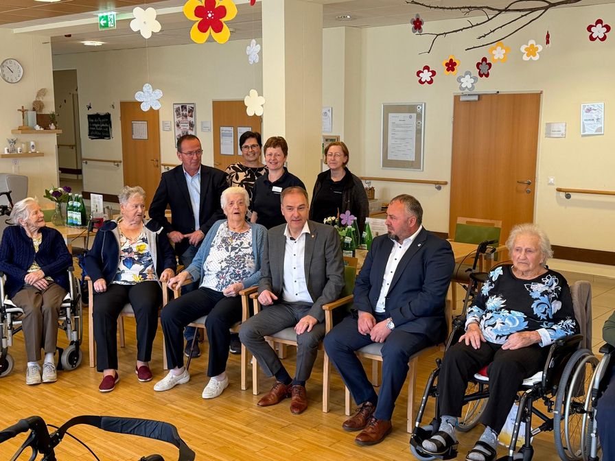 Eine Gruppe von Menschen sitzt in einem Wohnzimmer. Im Vordergrund sind mehrere ältere Personen in Rollstühlen und auf Stühlen zu sehen, während im Hintergrund einige Mitarbeitende stehen. Die Atmosphäre wirkt freundlich und einladend, mit Blumen-Dekorationen an den Wänden.